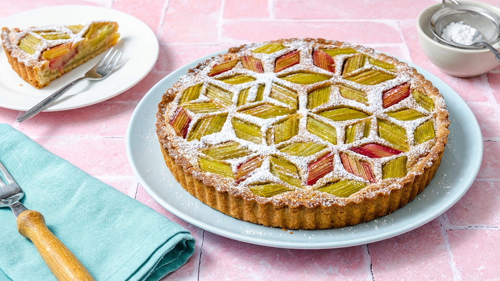 Ein runder Rhabarberkuchen mit einer geprägten Oberfläche aus Rhabarberstücken, bestäubt mit Puderzucker. Daneben befindet sich ein Stück des Kuchens auf einem weißen Teller, sowie ein blauer Servietten und ein Besteckset. Der Hintergrund ist rosa.