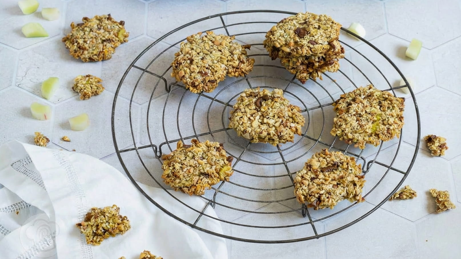 Auf einem runden Kuchengitter liegen mehrere haferflockenartige Kekse, die goldbraun gebacken sind. Einige kleine Bruchstücke sind um das Gestell verteilt. Die Keksoberfläche zeigt eine grobe Struktur mit Nüssen oder getrockneten Früchten.