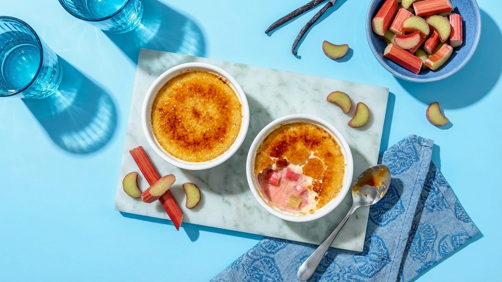 Zwei Schalen mit Crème Brûlée, eine mit Rhabarberstücken, stehen auf einem marmorierten Brett. Daneben sind einige Rhabarberstreifen und ein Löffel. Im Hintergrund sind Gläser mit Wasser und eine Schale mit Rhabarber zu sehen. Der Hintergrund ist hellblau.