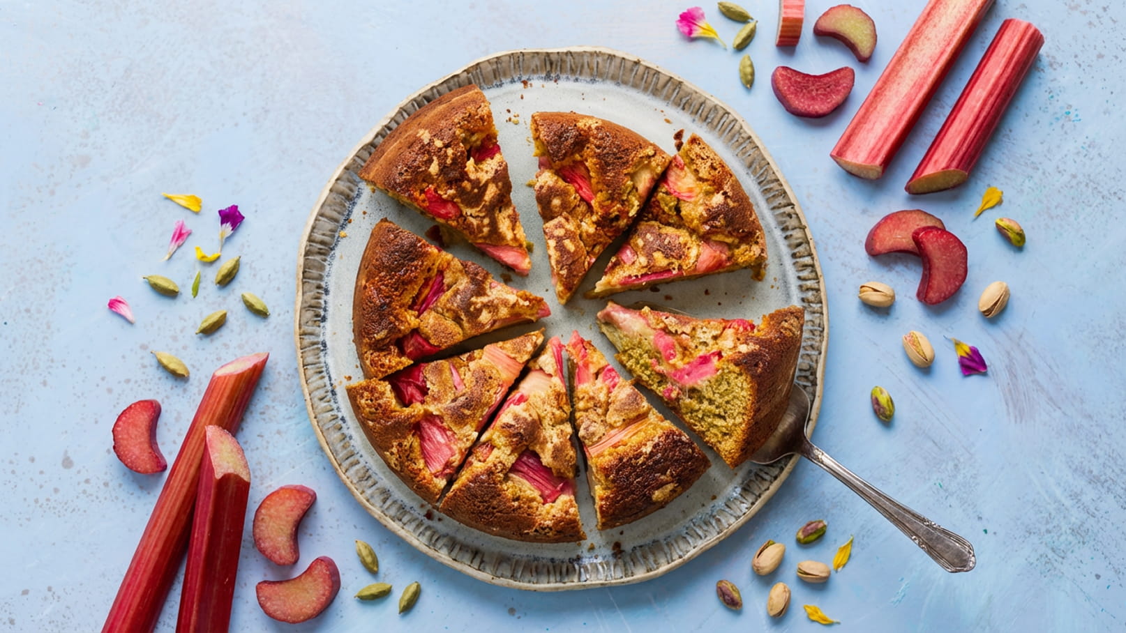 Ein runder Rhabarberkuchen in einem verzierter Teller, in acht gleichmäßige Stücke geschnitten. Um den Kuchen liegen frische Rhabarberstangen und einige Pistazien. Die Oberfläche des Kuchens ist goldbraun und teilweise mit Rhabarberstücken garniert.