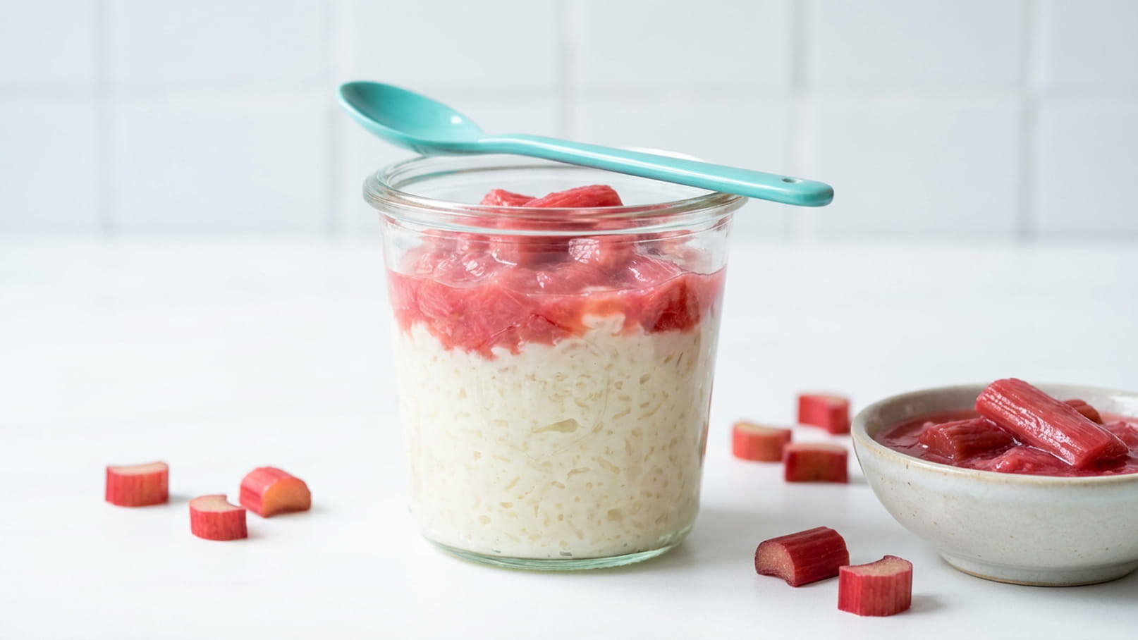 Ein Glas mit Milchreis, bedeckt mit stückigem Rhabarber, steht auf einem Tisch. Neben dem Glas liegt ein Löffel in Türkis. Auf dem Tisch befinden sich zusätzlich einige Rhabarberstücke und eine kleine Schüssel mit mehr Rhabarber.
