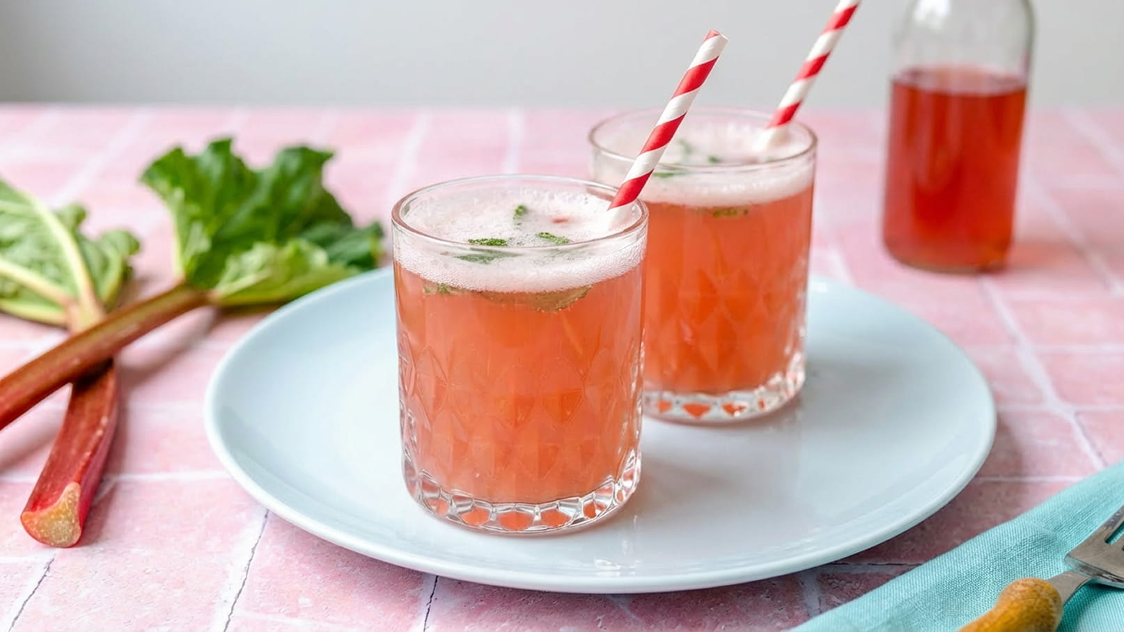 Zwei Gläser mit einem rosafarbenen Getränk stehen auf einem weißen Teller. Die Gläser haben gestreifte Trinkhalme und zeigen einen schaumigen Rand. Im Hintergrund ist eine Flasche und einige Rhabarberstängel sichtbar.