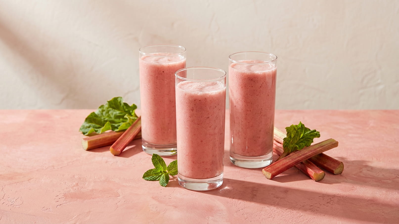 Drei hohe Gläser mit einem rosa Smoothie stehen auf einem rosa Tisch. Um die Gläser sind frische Rhabarberstängel und Minzblätter angeordnet. Die Szene vermittelt eine erfrischende und einladende Atmosphäre.