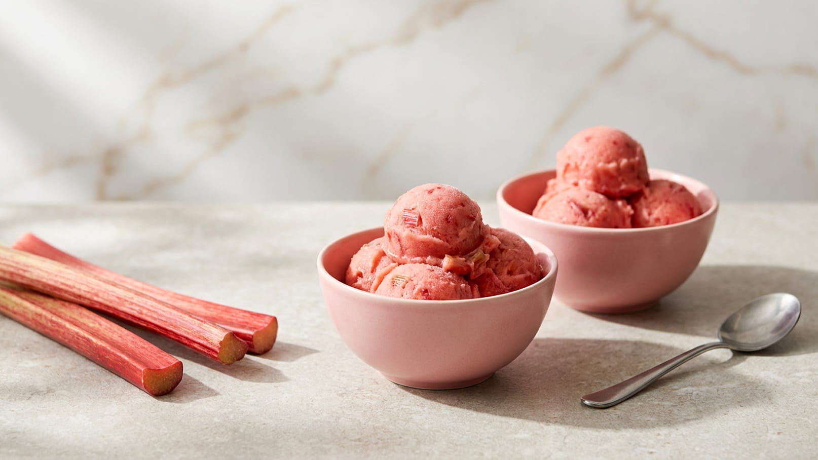 Zwei rosa Schalen mit Eis, gefüllt mit Erdbeereis, stehen auf einem Tisch. Neben den Schalen liegen einige Stangen Rhabarber. Ein Löffel befindet sich ebenfalls auf dem Tisch. Der Hintergrund zeigt eine sanfte, marmorierte Textur.