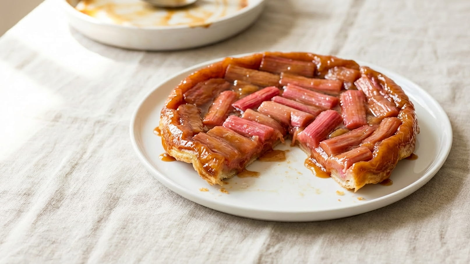 Eine Tarte Tatin mit Rhabarber auf einem weißen Teller. Die Torte hat eine glänzende, karamelisierte Oberseite und ist teilweise angeschnitten. Die Oberfläche zeigt gleichmäßig angeordnete Rhabarberstücke in rosa und rot. Der Hintergrund ist neutral und schlicht.