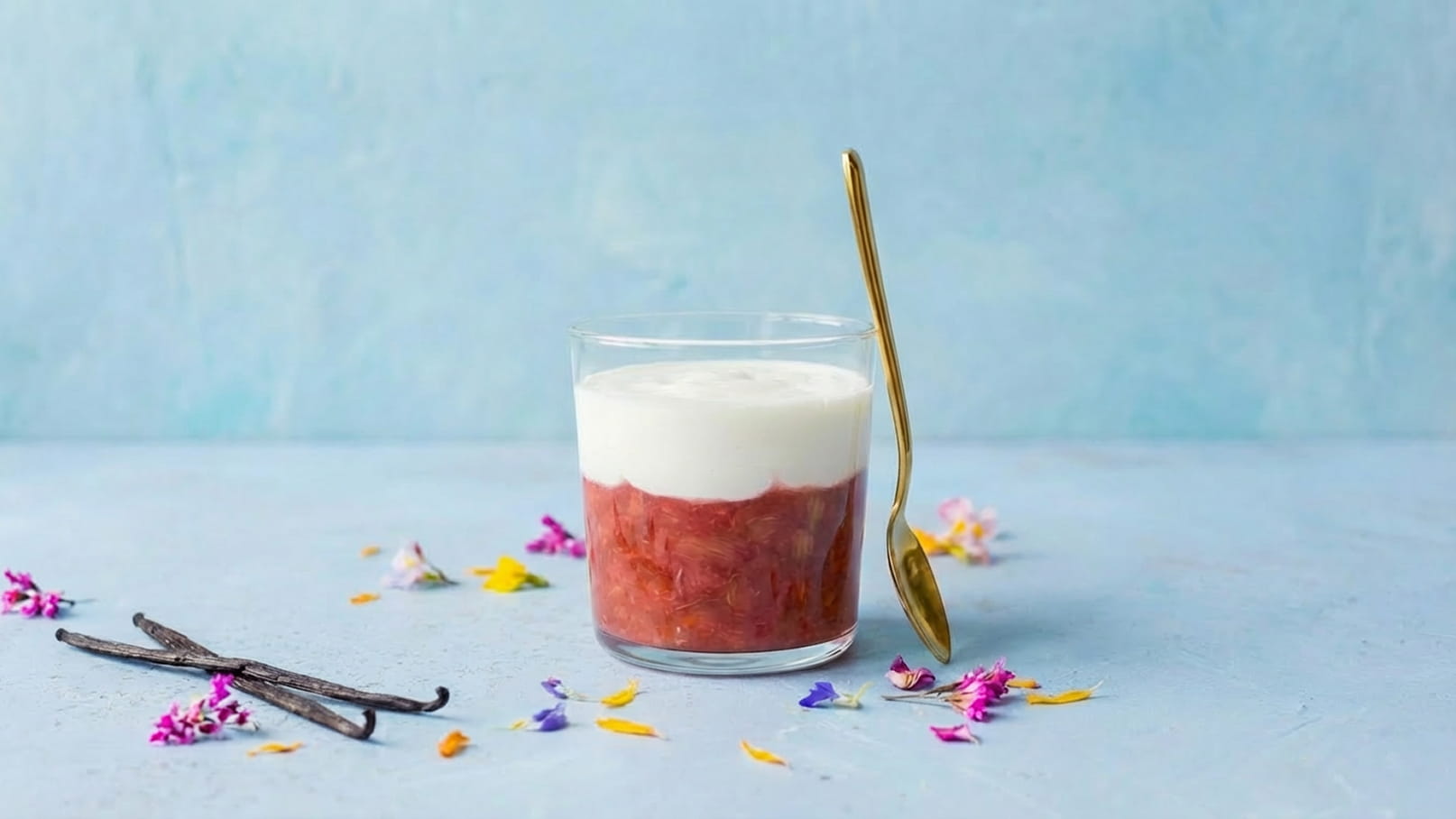 Ein Glas mit Rhabarberkompott, bedeckt mit einer Schicht Sahne. Neben dem Glas liegt ein goldener Löffel. Bunte essbare Blüten und Vanilleschoten sind auf der Tischoberfläche verteilt. Der Hintergrund ist sanft blau.