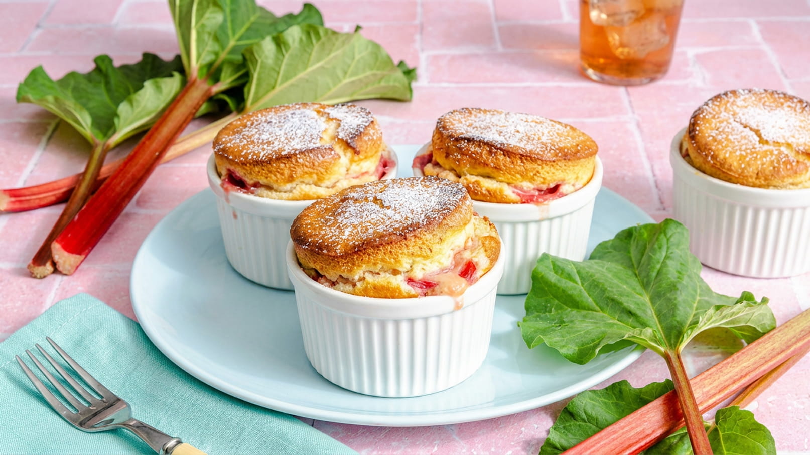 Vier kleine Aufläufe mit Rhabarber in weißen Förmchen, bestäubt mit Puderzucker, stehen auf einem hellblauen Teller. Umgeben sind sie von frischen Rhabarberstangen und grünen Blättern. Im Hintergrund ist ein Getränk in einem Glas sichtbar.