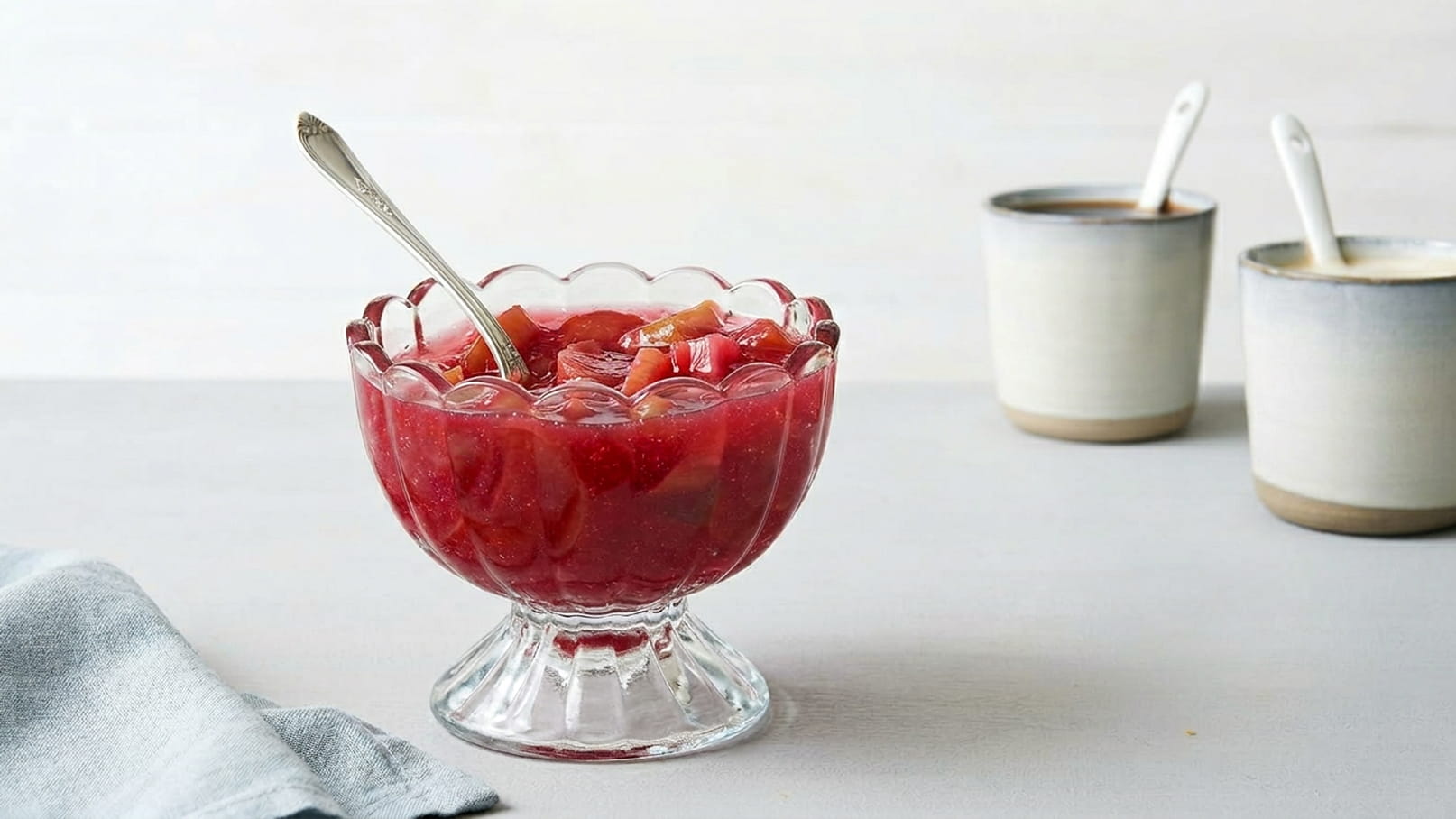 Eine Schüssel aus Glas enthält eine rote Fruchtgelee-Mischung mit Stücken von Früchten. Ein Löffel steckt in der Gelee. Im Hintergrund sind zwei Tassen mit Löffeln zu sehen.