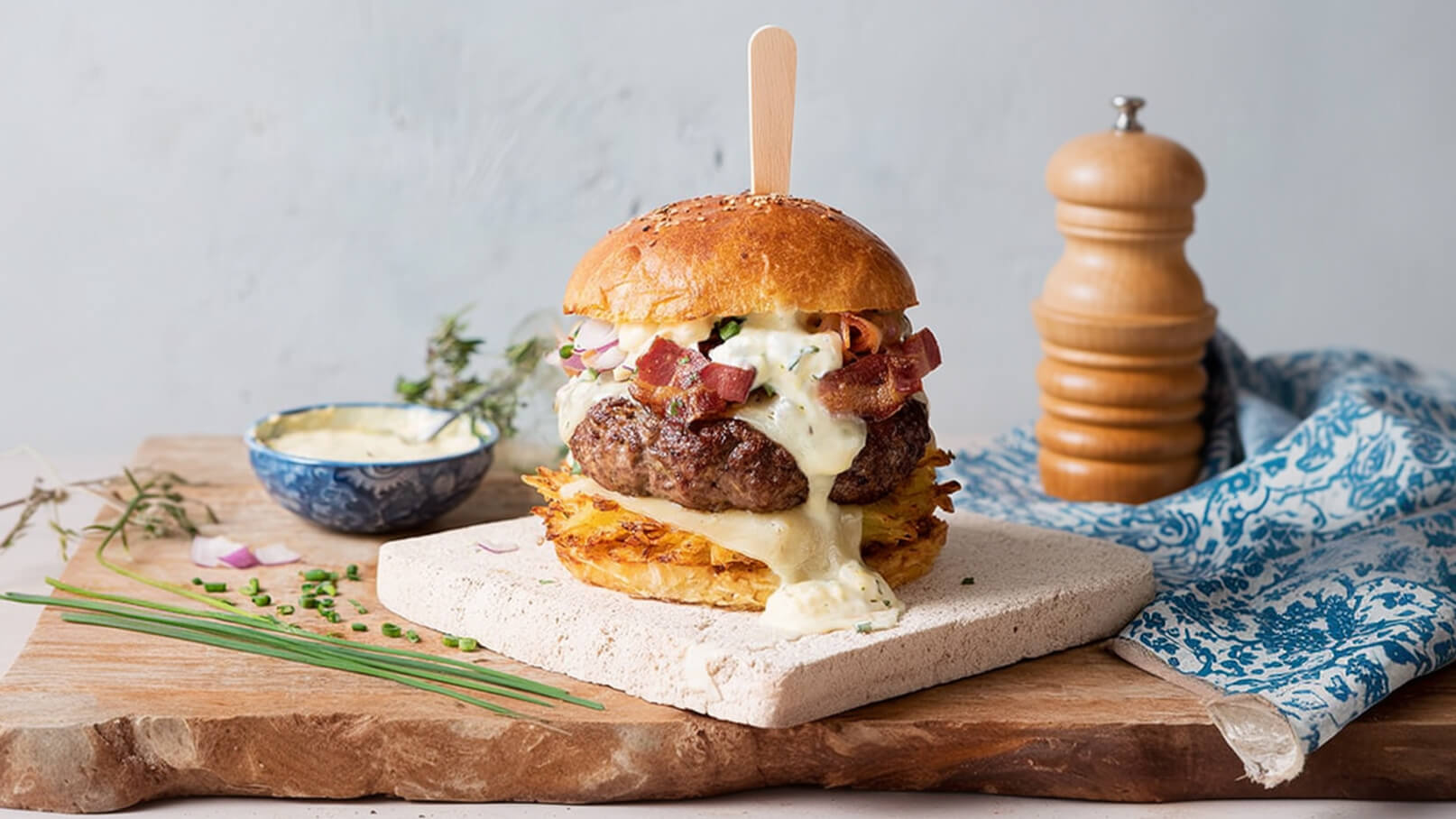 Ein üppiger Burger auf einem Holzbrett, gefüllt mit einem saftigen Patty, geschmolzenem Käse, Speck und frischen Zutaten. Eine kleine Schüssel mit Sauce befindet sich neben dem Burger. Dekorative Kräuter und Frühlingszwiebeln liegen auf dem Brett.