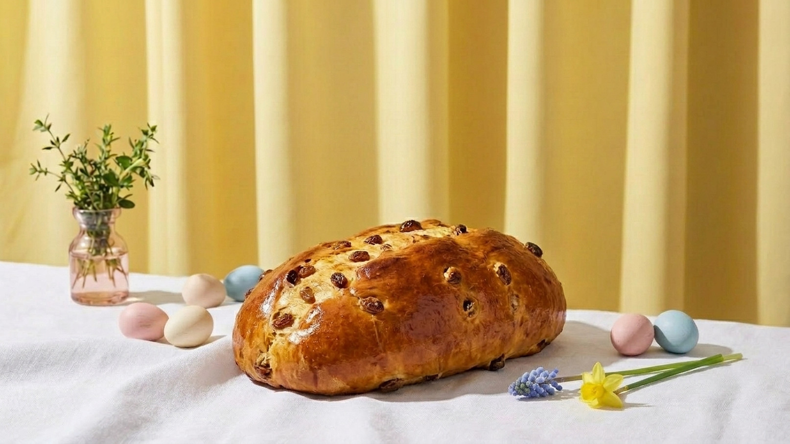 Ein frisch gebackener, goldbrauner Hefezopf mit Rosinen liegt auf einem weißen Tisch. Rund um ihn sind bunte Eier verteilt, und daneben steht ein kleines Glas mit frischen Kräutern. Der Hintergrund ist gelb und hell.