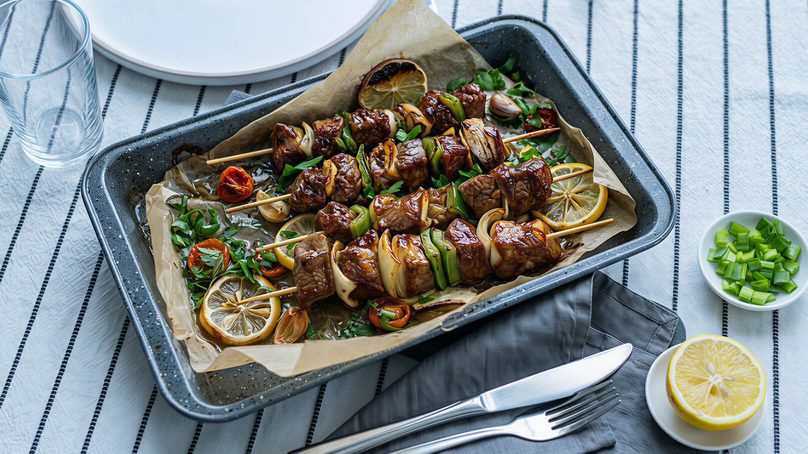 Gegrillte Spieße mit Fleisch, Gemüse und Zitronenscheiben auf einem Backblech, umgeben von frischen Kräutern. Serviert auf einem Tisch mit Besteck, einem Glas und einem weiteren Teller im Hintergrund. Ein weiteres Gericht mit geschnittenen Frühlingszwiebeln und einer Zitrone ist daneben platziert.
