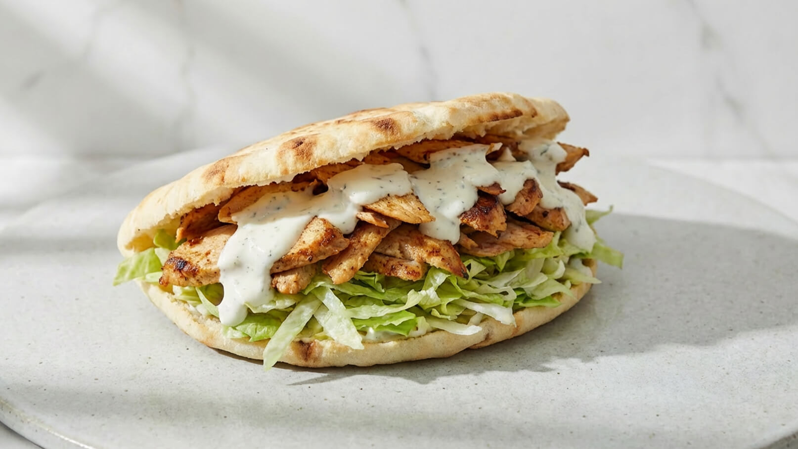 Eine frische Fladenbrot-Sandwich gefüllt mit gegrilltem Hähnchen, knackigem Salat und einer weißen Sauce. Das Fladenbrot ist leicht gebräunt und die Füllung ist großzügig geschichtet, was eine appetitliche und harmonische Mahlzeit verspricht.