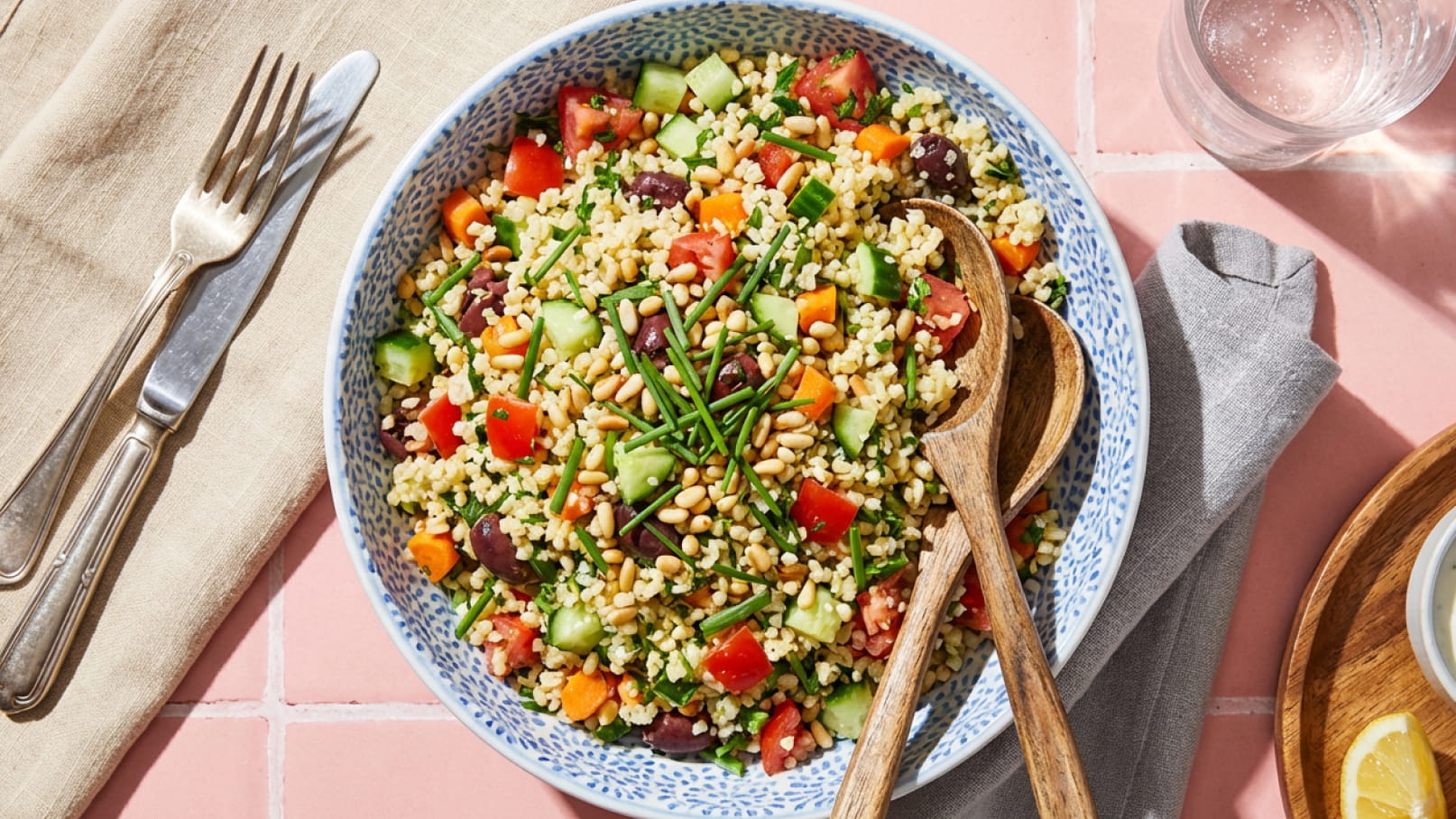 Eine große Schüssel mit buntem Couscous-Salat, der aus verschiedenen Gemüsesorten wie Tomaten und Gurken, sowie Pinienkernen und frischen Kräutern besteht. Holzlöffel liegen neben der Schüssel, die auf einem rosa Tisch gedeckt ist.