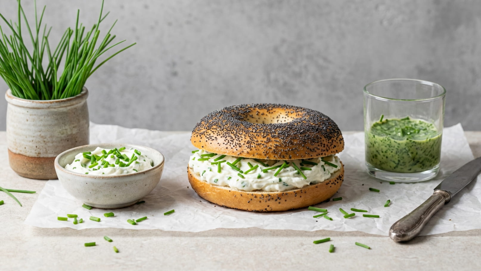 Ein Sandwich aus einem Mohnbagel mit einer großzügigen Schicht Kräutercreme, garniert mit frischem Schnittlauch. Daneben befindet sich eine kleine Schüssel mit Kräutercreme und ein Glas mit einer grünen Sauce. Im Hintergrund steht ein kleiner Topf mit frischem Schnittlauch.