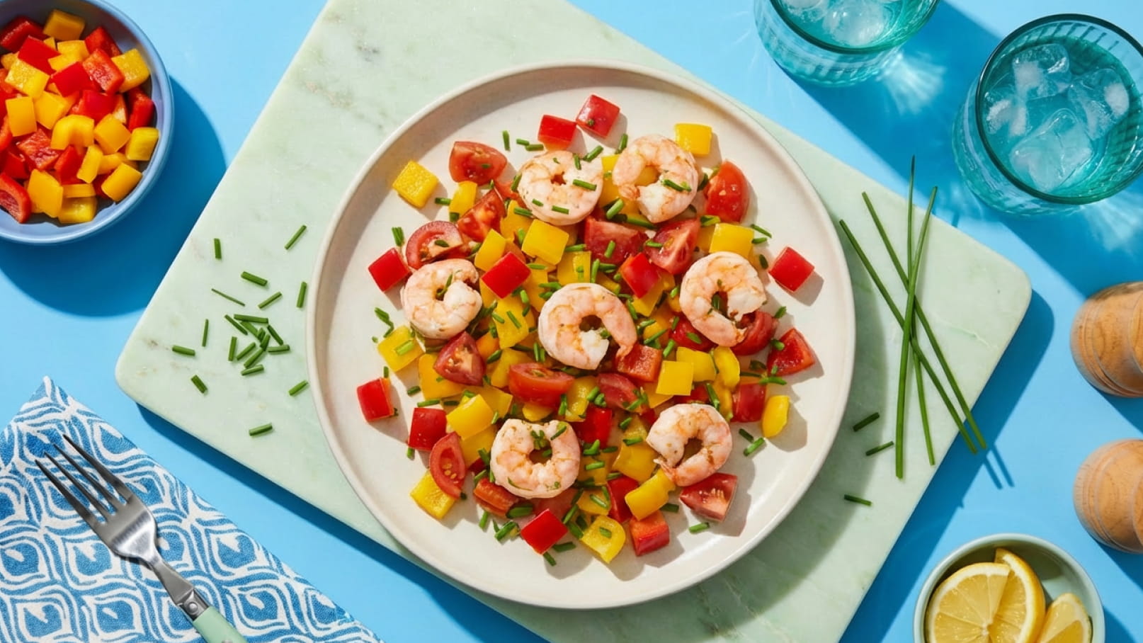 Ein Teller mit Garnelen, bunten Paprika-Streifen, Tomaten und frischem Schnittlauch. Neben dem Teller befindet sich eine Schüssel mit gewürfelten Mangos und Paprika sowie zwei Gläser mit Wasser. Der Hintergrund ist hellblau.