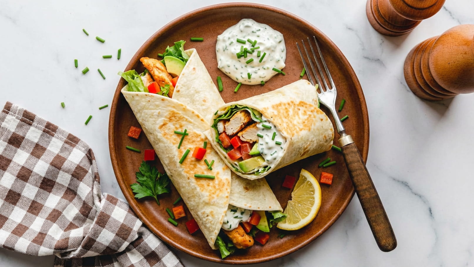 Ein Teller mit zwei gefüllten Tortilla-Wraps, die mit Hähnchen, Salat, Paprika und Gurke belegt sind. Daneben befindet sich ein Dip aus Joghurt und Kräutern, sowie eine Zitronenscheibe. Ein kariertes Geschirrtuch und Holzsalz- und Pfeffermühlen sind ebenfalls sichtbar.