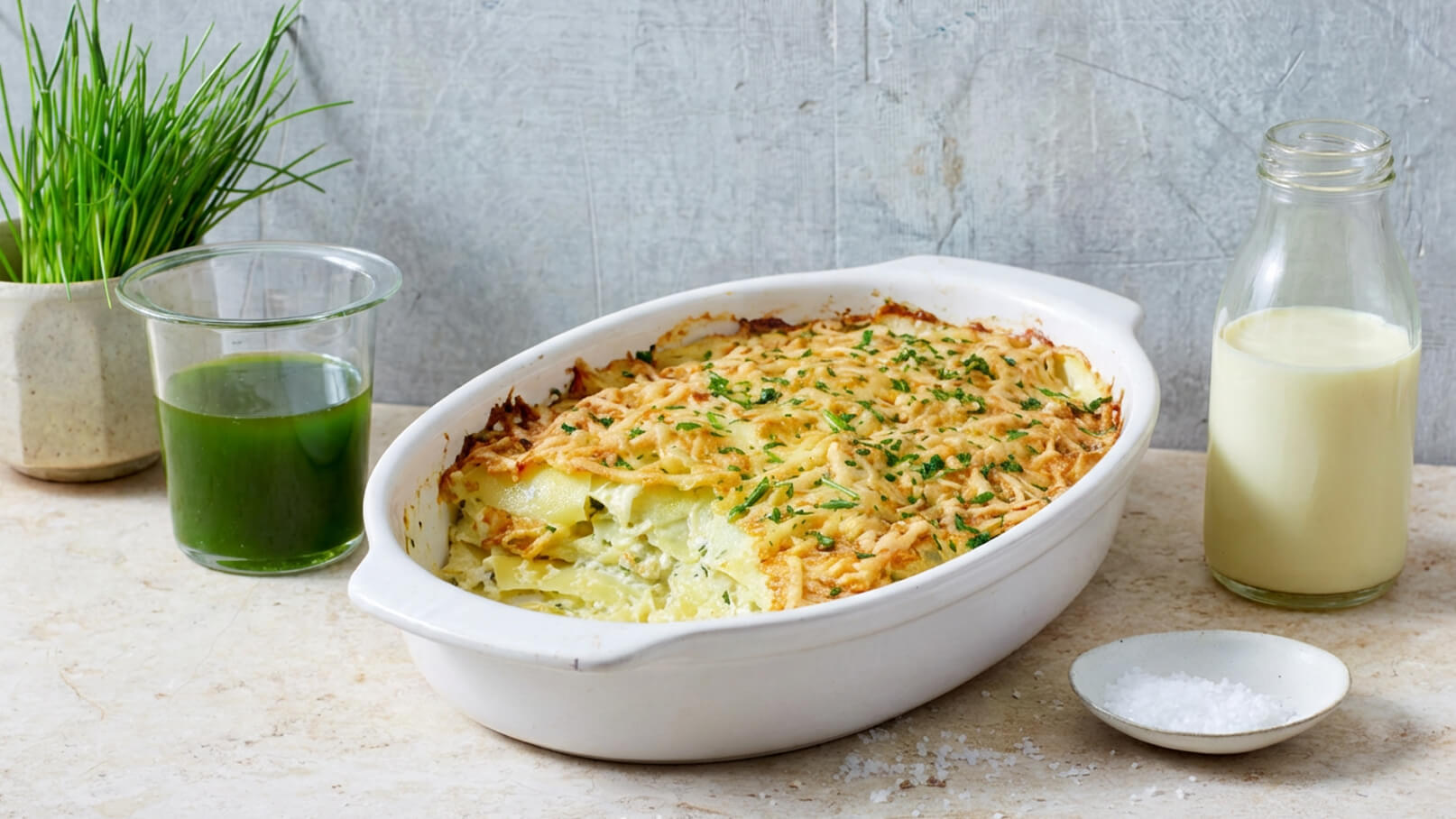 Ein Auflaufform mit einem goldenen Kartoffelgericht, garniert mit frischem Schnittlauch. Daneben stehen ein Glas grüner Saft, eine Flasche Milch und eine kleine Schale mit Salz. Der Hintergrund ist schlicht und hell.