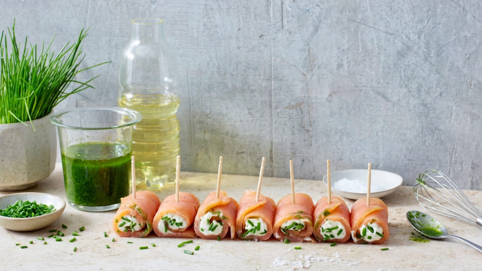 Das Bild zeigt eine Reihe von mit Frischkäse und Schnittlauch gefüllten Schinkenröllchen, die auf einem Tisch angerichtet sind. Neben den Röllchen stehen ein Glas mit grünem Dressing, Öl und frische Schnittlauchzweige.