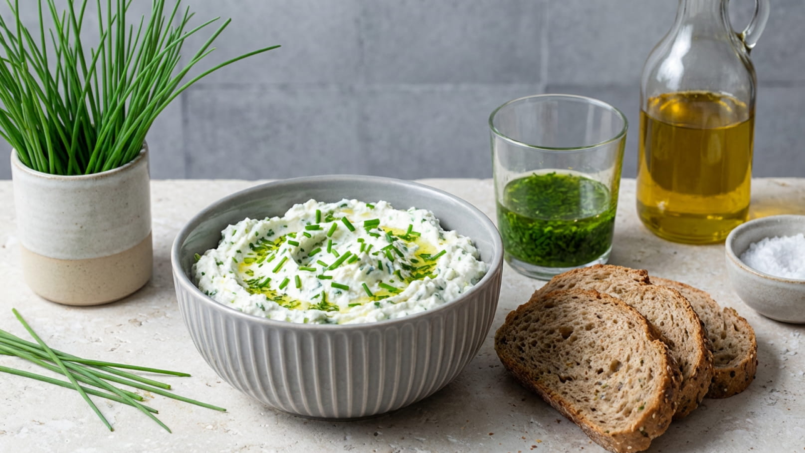 Eine Schüssel mit Kräuterquark, garniert mit frischem Schnittlauch, steht auf einem Tisch. Daneben liegen einige Scheiben Vollkornbrot, ein Glas mit grüner Soße, eine Flasche Olivenöl und eine Schale mit Salz. Frische Schnittlauchhalme ergänzen die Präsentation.
