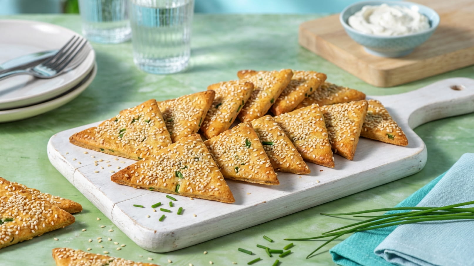 Ein Holzbrett mit dreieckigen Keksen, bestreut mit Sesam und Schnittlauch. Im Hintergrund sind zwei Gläser Wasser und eine Schüssel mit Creme zu sehen. Die Kekse sind knusprig und ansprechend angerichtet, passend zu einem gemütlichen Snackmoment.