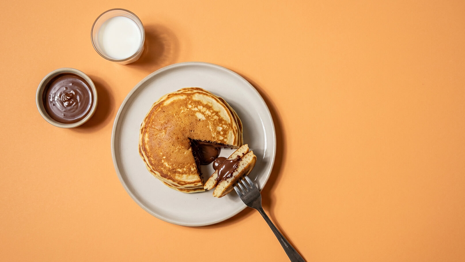 Ein Teller mit stapelweisen Pfannkuchen, aus denen ein schokoladiger Inhalt herausquillt. Daneben ein Glas Milch und eine Schüssel mit Schokoladensoße. Der Hintergrund ist in einem warmen Orange gehalten.