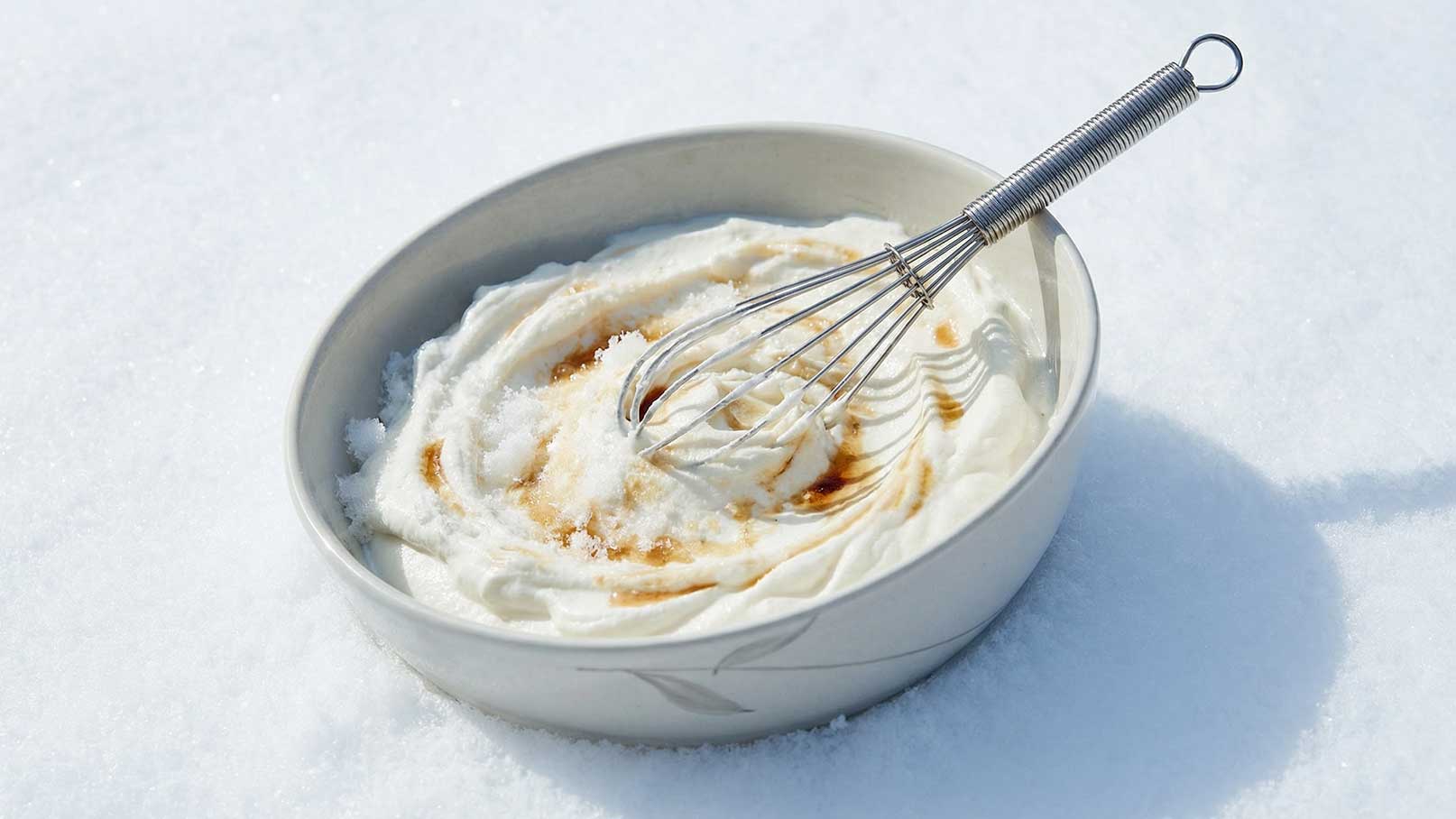 Eine Schüssel mit cremiger, weißer Masse, vermutlich Sahne oder Joghurt, ist in einer schneebedeckten Umgebung platziert. Ein Schneebesen steckt in der Mischung, die mit einer Flüssigkeit, möglicherweise Sirup, verfeinert ist.