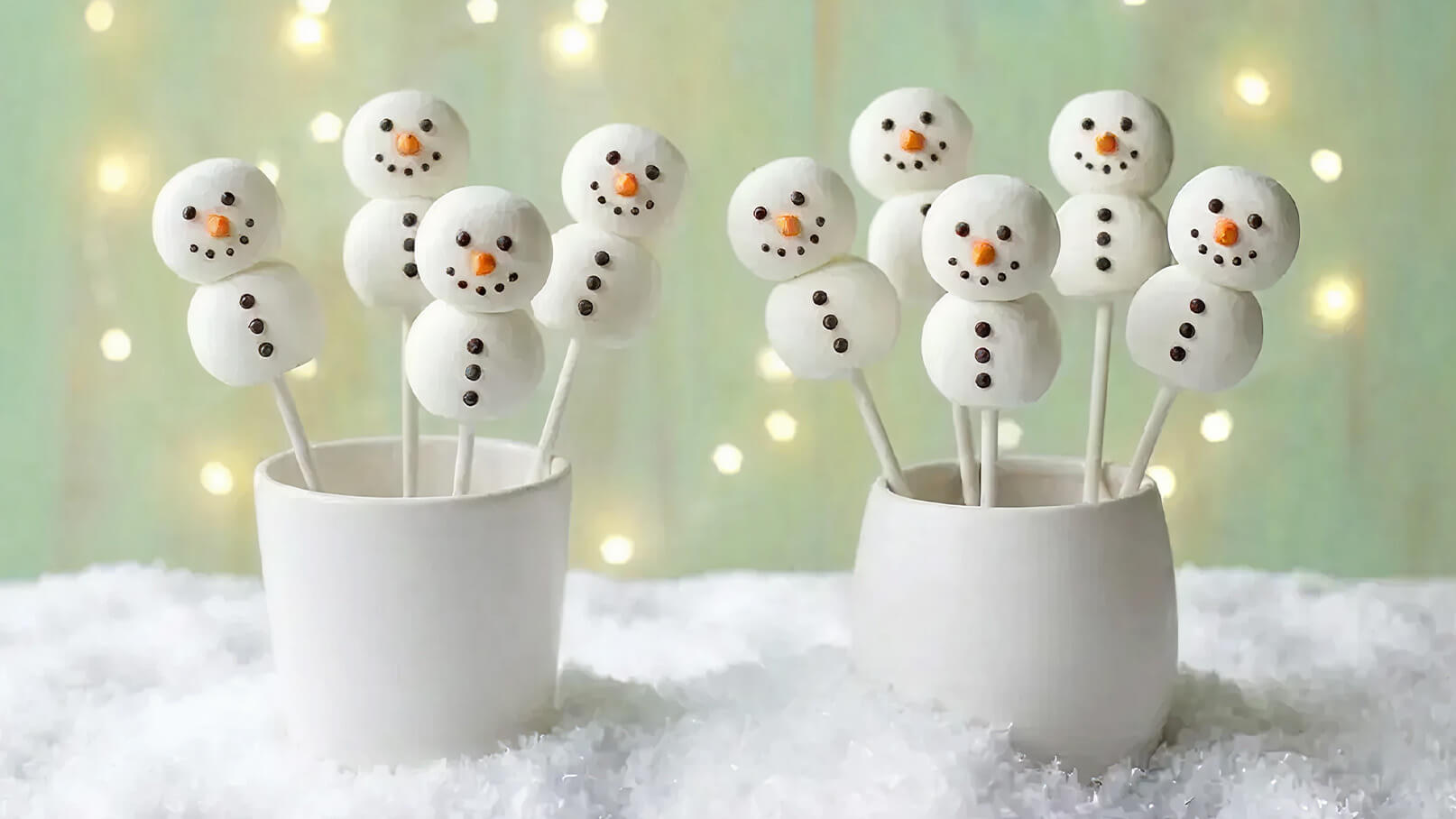 Zwei Töpfe enthalten festlich dekorierte Schneemann-Lollis aus weißem süßem Material. Die Schneemänner haben orangefarbene Karottennasen und schwarze Zuckerperlen als Augen und Knöpfe. Im Hintergrund glitzert dezentes Licht. Die Lollis stehen in einer schneebedeckten Umgebung.