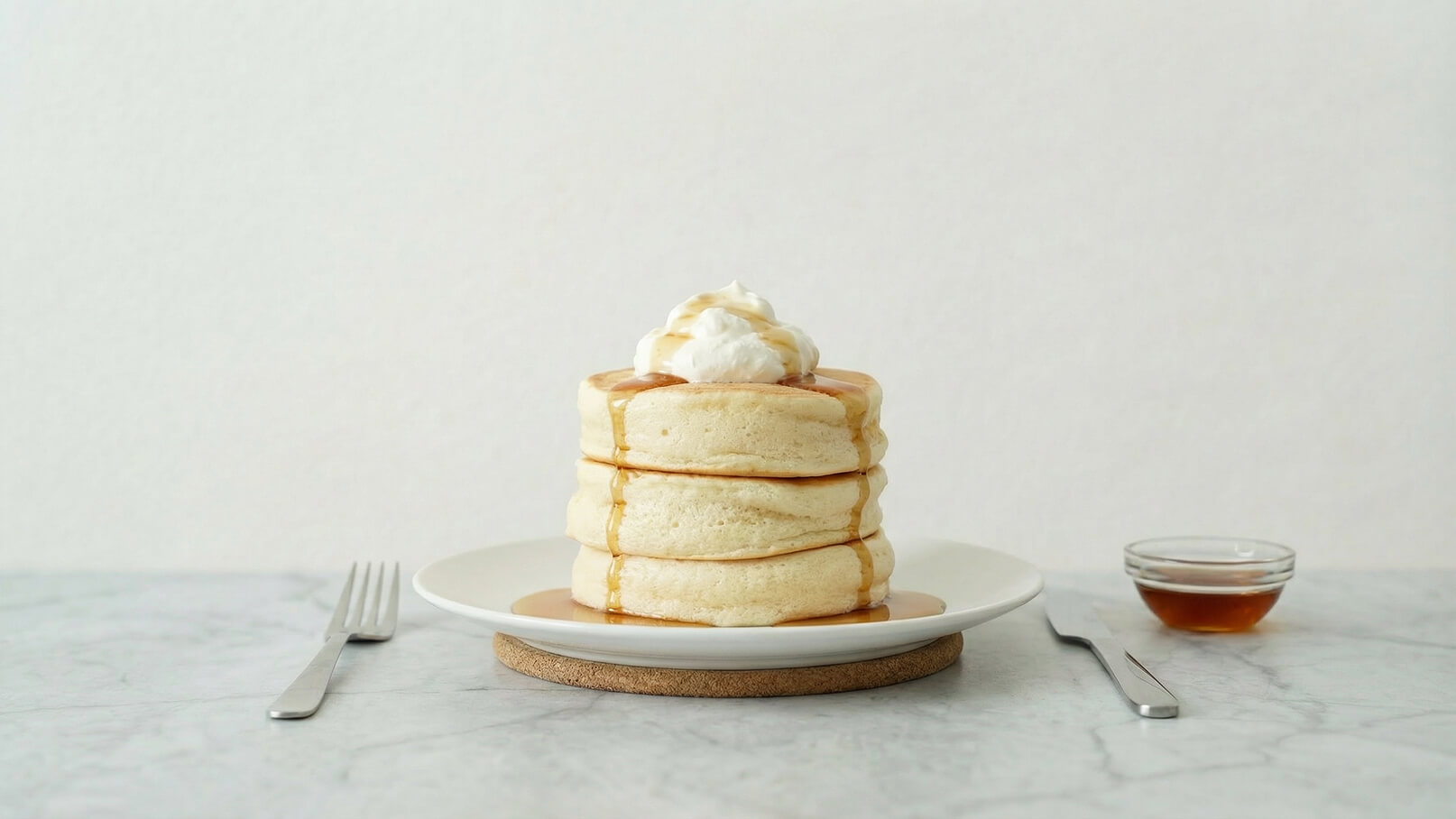 Drei dicke Pancakes übereinandergestapelt, mit einem Klecks Sahne und einem Schuss Sirup darauf. Sie befinden sich auf einem weißen Teller auf einem Untersetzer. Neben dem Teller steht eine kleine Schale mit etwas Sirup. Der Hintergrund ist neutral und hell.