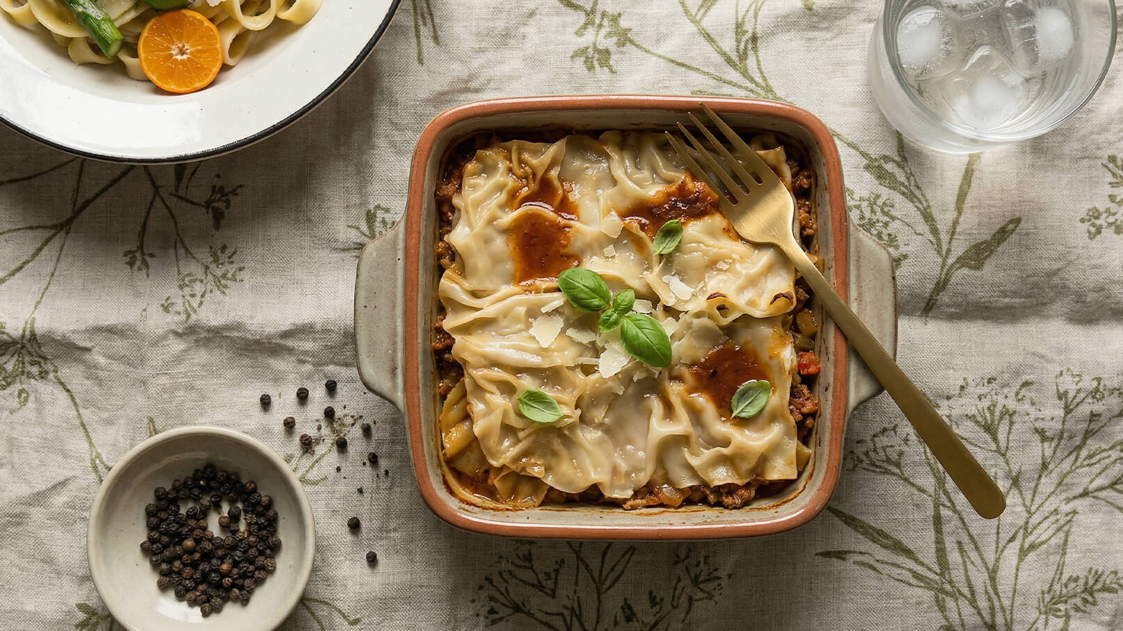 Eine Auflaufform mit Lasagne, garniert mit frischem Basilikum. Daneben befindet sich eine kleine Schale mit Pfefferkörnern und ein Glas Wasser. Im Hintergrund ist ein Teller mit Pasta und einer Orange. Der Tisch ist mit einer naturmusternden Tischdecke dekoriert.