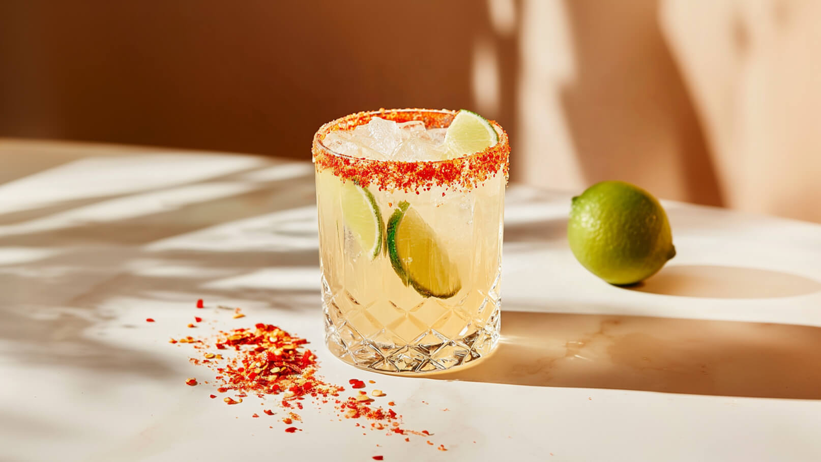 Ein Glas mit einem erfrischenden Cocktail gefüllt mit Eiswürfeln und Limettenscheiben. Der Glasrand ist mit einer roten Gewürzmischung verziert. Neben dem Cocktail liegt eine frische Limette auf dem Tisch.