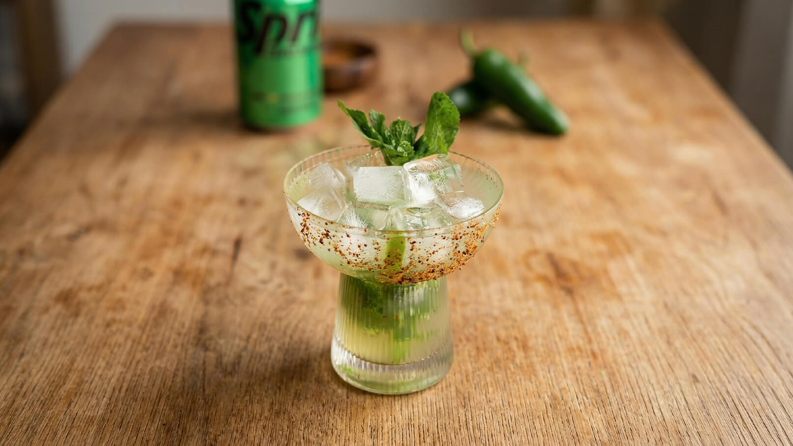 Ein Cocktailglas mit einem Getränk, gefüllt mit Eiswürfeln und frischen Minzeblättern. Um den Glasrand ist eine rote Gewürzschicht sichtbar. Im Hintergrund stehen eine grüne Dose und eine Jalapeño. Die Holzoberfläche vermittelt eine natürliche Atmosphäre.