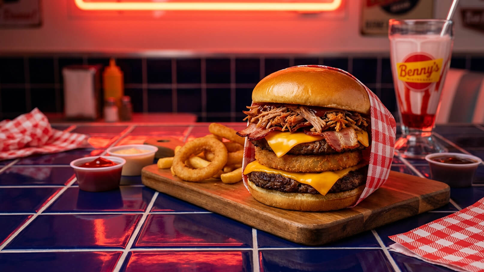 Ein großer Burger auf einem Holzbrett, geschichtet mit zwei Cheeseburger-Patties, knusprigem Speck, Pulled Pork und einem Brötchen. Daneben liegen frittierte Zwiebelringe und zwei kleine Schüsseln mit Ketchup und einer anderen Sauce. Im Hintergrund steht ein Glas mit einem Milchshake.