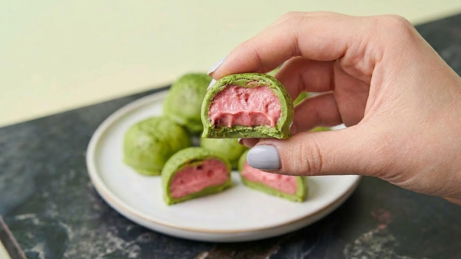 Eine Hand hält ein halbiertes Dessert mit grüner Außenhülle und pinker Füllung. Auf einem Teller im Hintergrund liegen mehrere dieser Desserts. Die Farben und die Form deuten auf eine süße Leckerei hin.