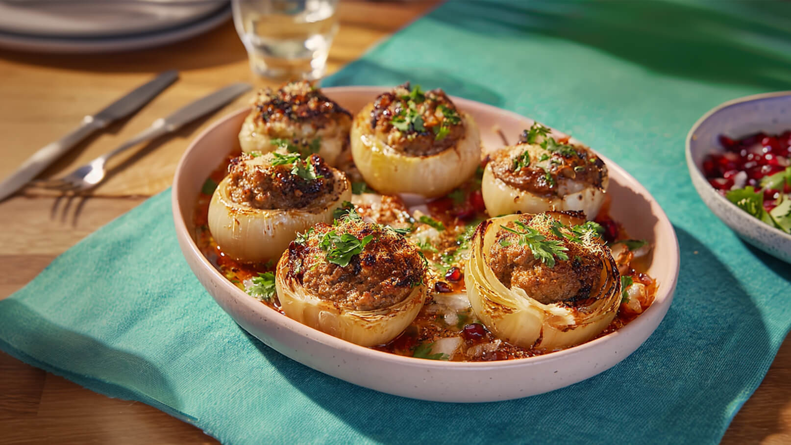 Ein Teller mit gefüllten Zwiebeln, die eine herzhafte Füllung und frische Kräuter obendrauf haben. Die Zwiebeln sind goldbraun gebacken und liegen in einer leicht würzigen Sauce. Daneben befindet sich eine kleine Schüssel mit Granatapfelkernen und einer Kräutermischung.