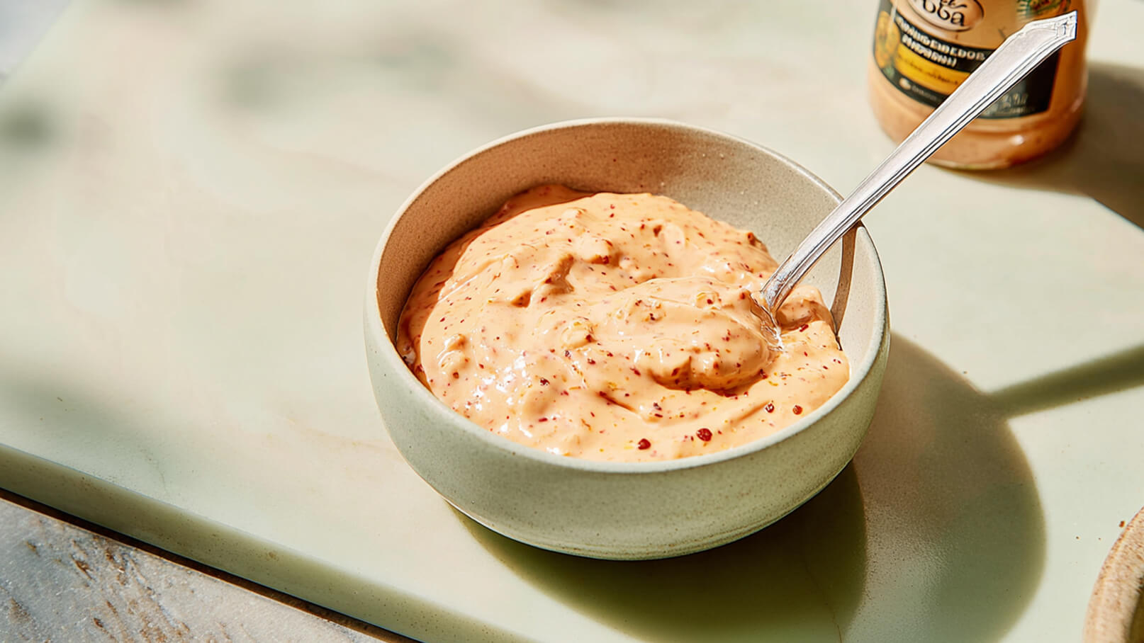 Eine helle, cremige Sauce mit rötlichen Sprenkeln befindet sich in einer runden Schüssel. Ein Löffel liegt in der Sauce. Im Hintergrund ist ein Glas mit einer ähnlichen Sauce sichtbar.