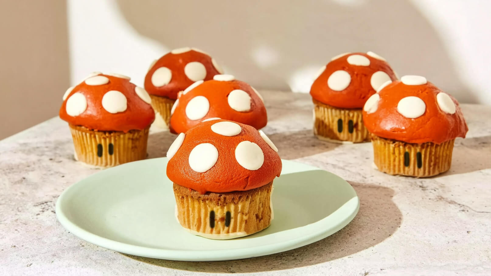 Sechs Cupcakes auf einem Tisch, die wie Pilze gestaltet sind. Der Cupcake auf einem hellgrünen Teller hat einen roten Hut mit weißen Punkten. Die anderen Cupcakes im Hintergrund haben ähnliche Farben und Designs.
