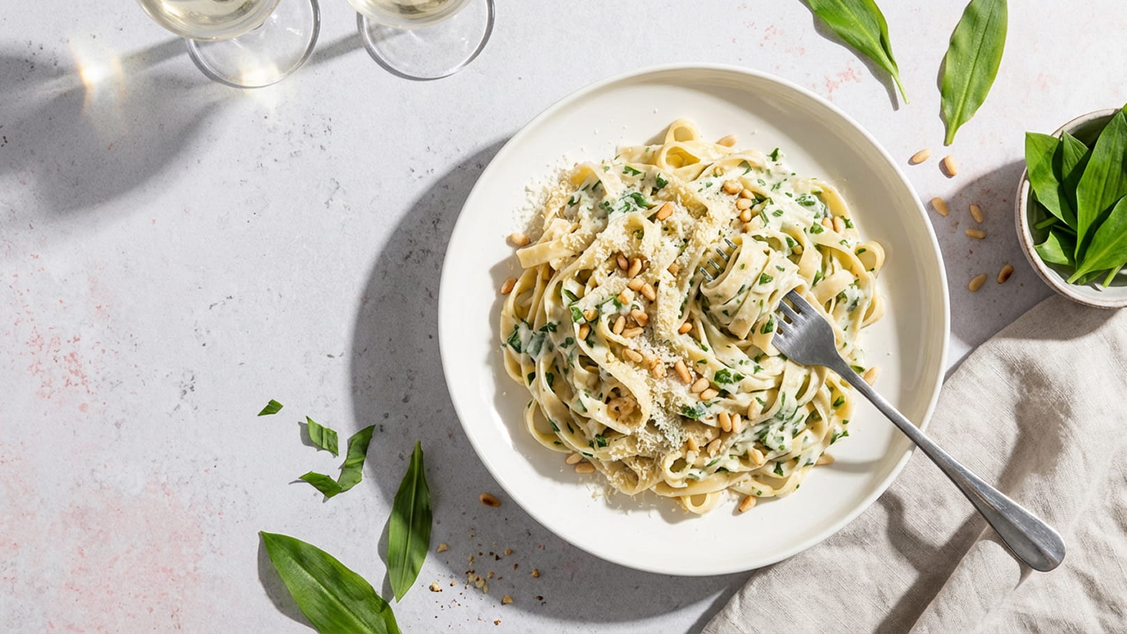 Ein Weißer Teller mit cremigen Tagliatelle, garniert mit frischen Kräutern und Pinienkernen. Im Hintergrund sind zwei Gläser Weißwein sichtbar sowie einige Blätter, die das Gericht umgeben.