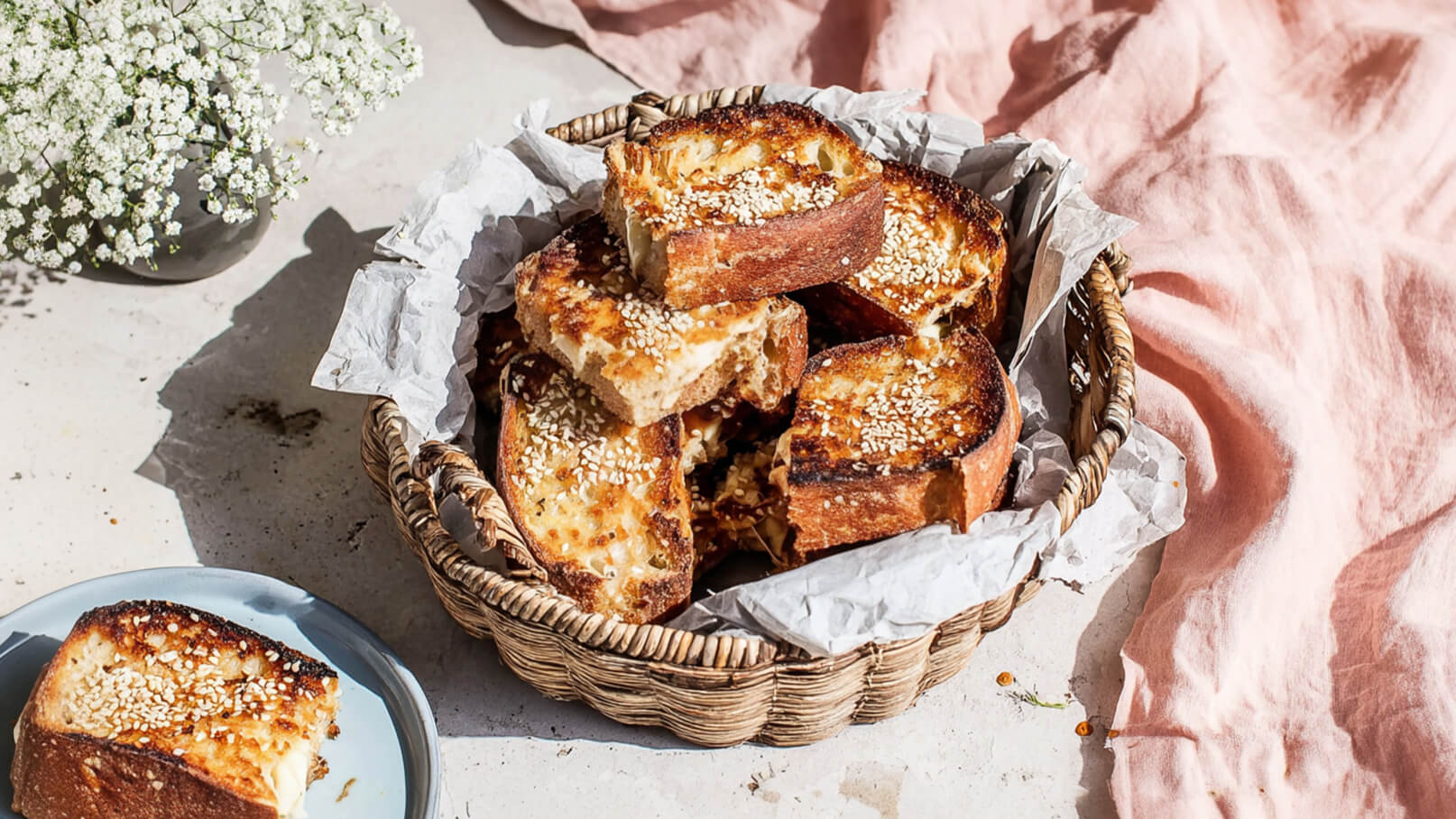 Ein Korb voller gerösteter Brotscheiben, mit Sesam bestreut, liegt auf crumpled Papier. Das Brot hat eine goldbraune Farbe und ist leicht knusprig. Im Hintergrund ist ein zarter Blumenstrauß zu sehen, der die Szene zusätzlich belebt.