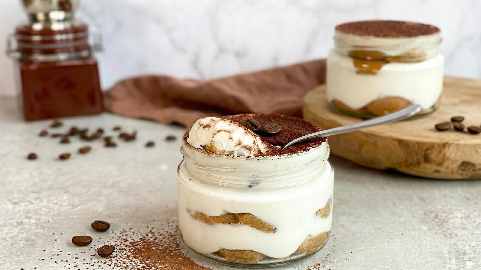 Zwei Gläser mit Tiramisu sind auf einer Tischoberfläche platziert. Ein Glas enthält abwechselnde Schichten aus Mascarpone-Creme und Löffelbiskuits, bestäubt mit Kakaopulver. Ein Esslöffel befindet sich im Glas. Kaffeebohnen und ein Kaffeebereiter sind im Hintergrund sichtbar.