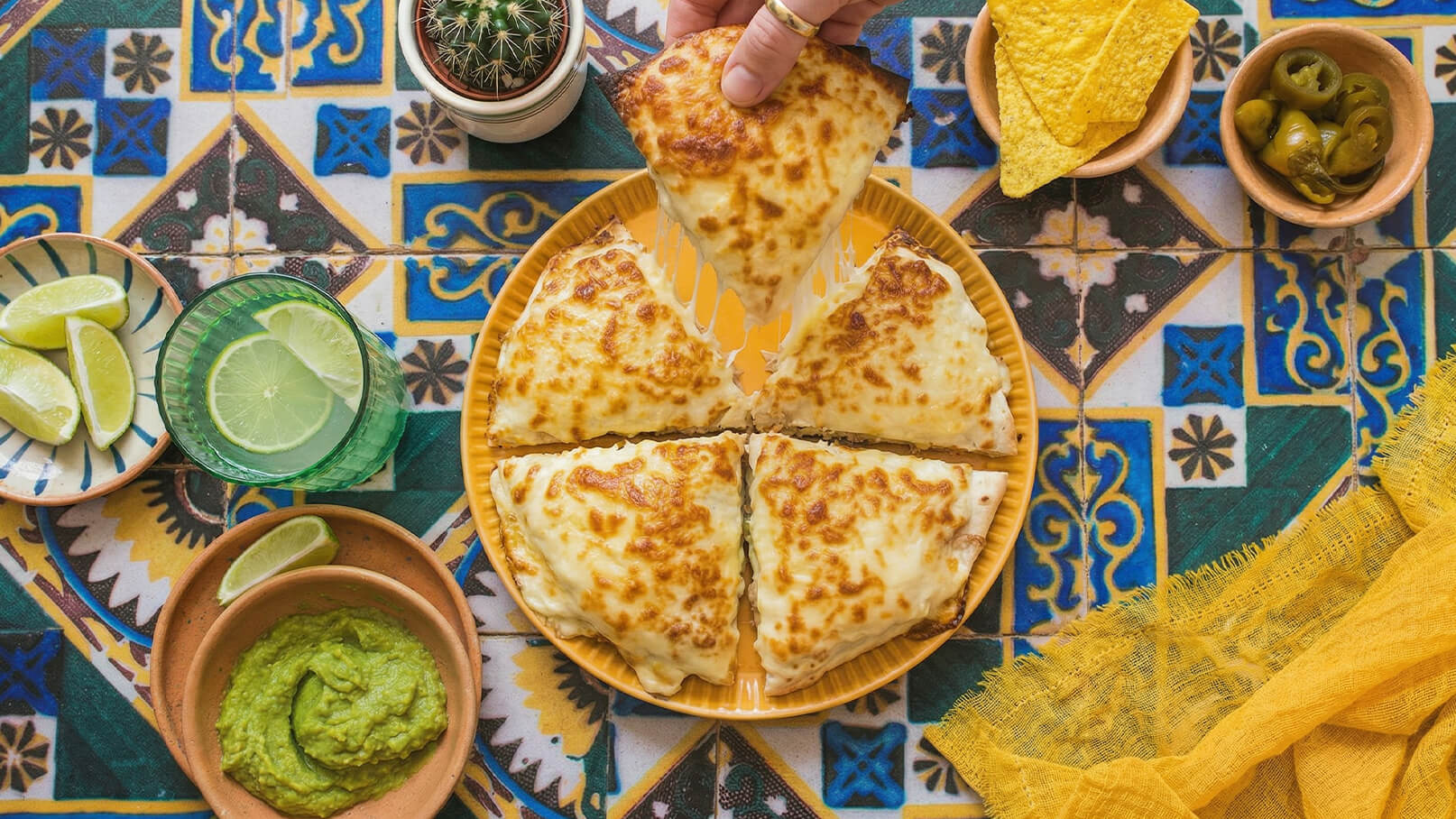 Eine Person hebt ein Stück Pizza von einem runden Teller, der in vier gleichgroße Stücke geschnitten ist. Um den Teller liegen Limetten, Guacamole, Tortilla-Chips und Oliven. Der Tisch ist mit einem bunten, gemusterten Tuch bedeckt.