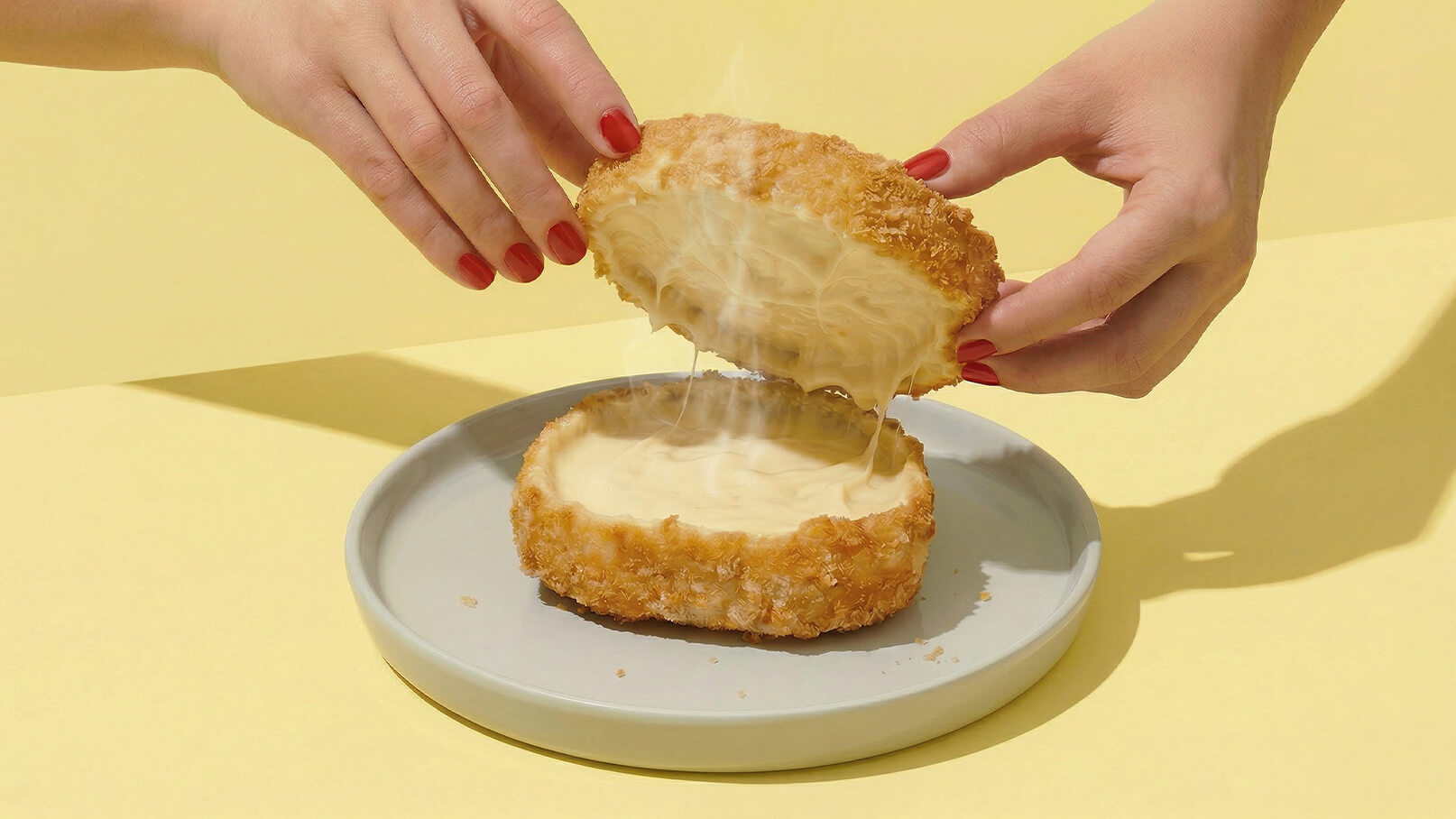 Zwei Hände heben den oberen Teil eines panierten Käsekuchens an. Der Käsekuchen zeigt eine cremige Füllung, die beim Öffnen herauszieht. Der Kuchen liegt auf einem grauen Teller vor einem gelben Hintergrund.