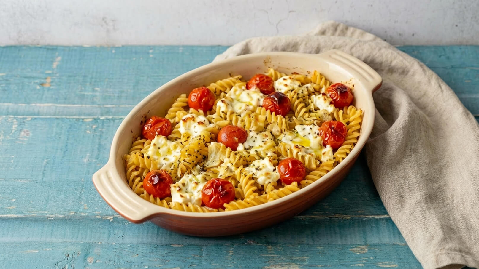 Ein Auflauf mit Spiralnudeln, überzogen mit einer cremigen Sauce und garniert mit gerösteten Kirschtomaten. Der Auflauf ist in einer ovalen Schale serviert und steht auf einem blauen Holzuntergrund, begleitet von einem grauen Küchentuch.