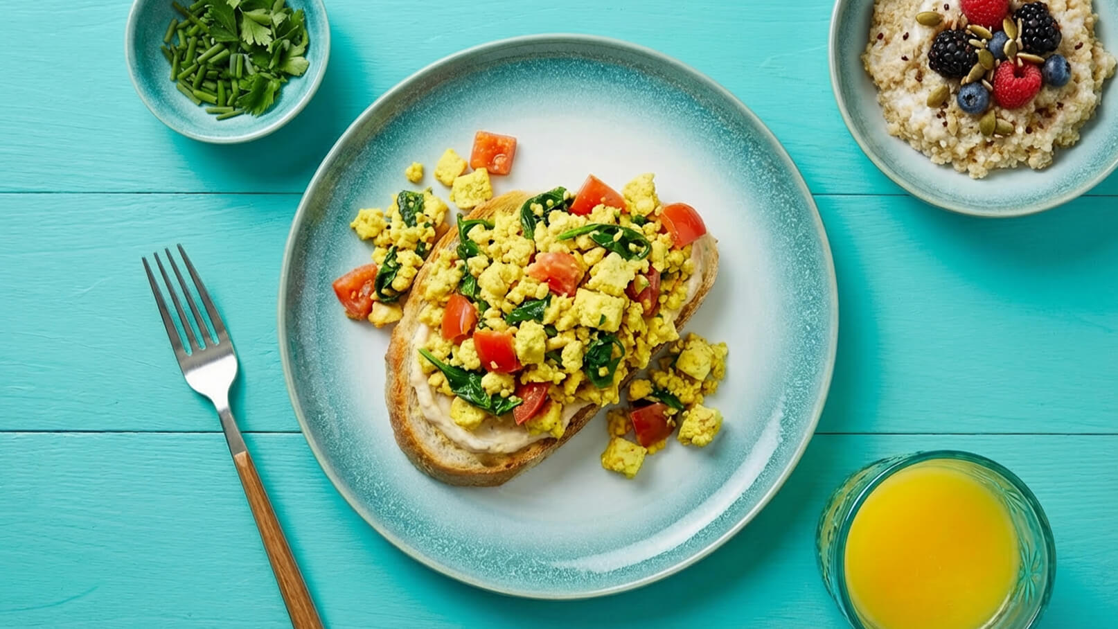 Ein Teller mit geröstetem Brot, belegt mit Rührei aus Tofu, Spinat und Tomaten. Daneben eine Schüssel mit Haferbrei und frischen Beeren sowie ein Glas Orangensaft. Der Hintergrund ist in einem hellen Blau gehalten.