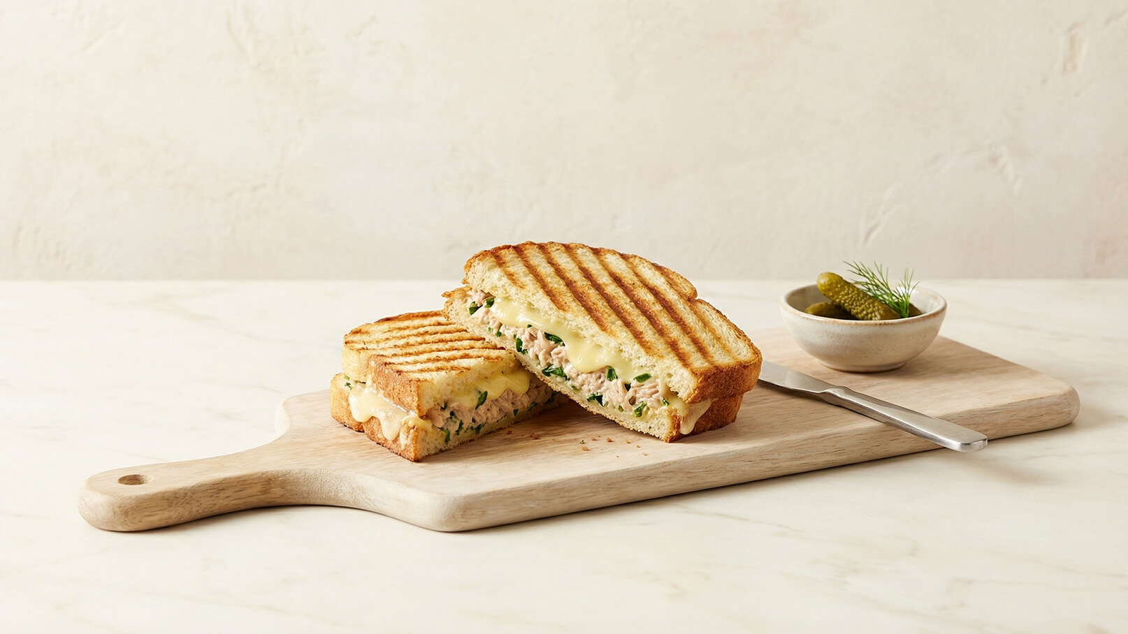 Ein gegrilltes Sandwich mit einer Füllung aus Fisch und Kräutern, auf einem Holzbrett präsentiert. Es ist in zwei Hälften geschnitten, die Grillstreifen sind sichtbar. Eine Schale mit eingelegten Gewürzgurken liegt daneben.