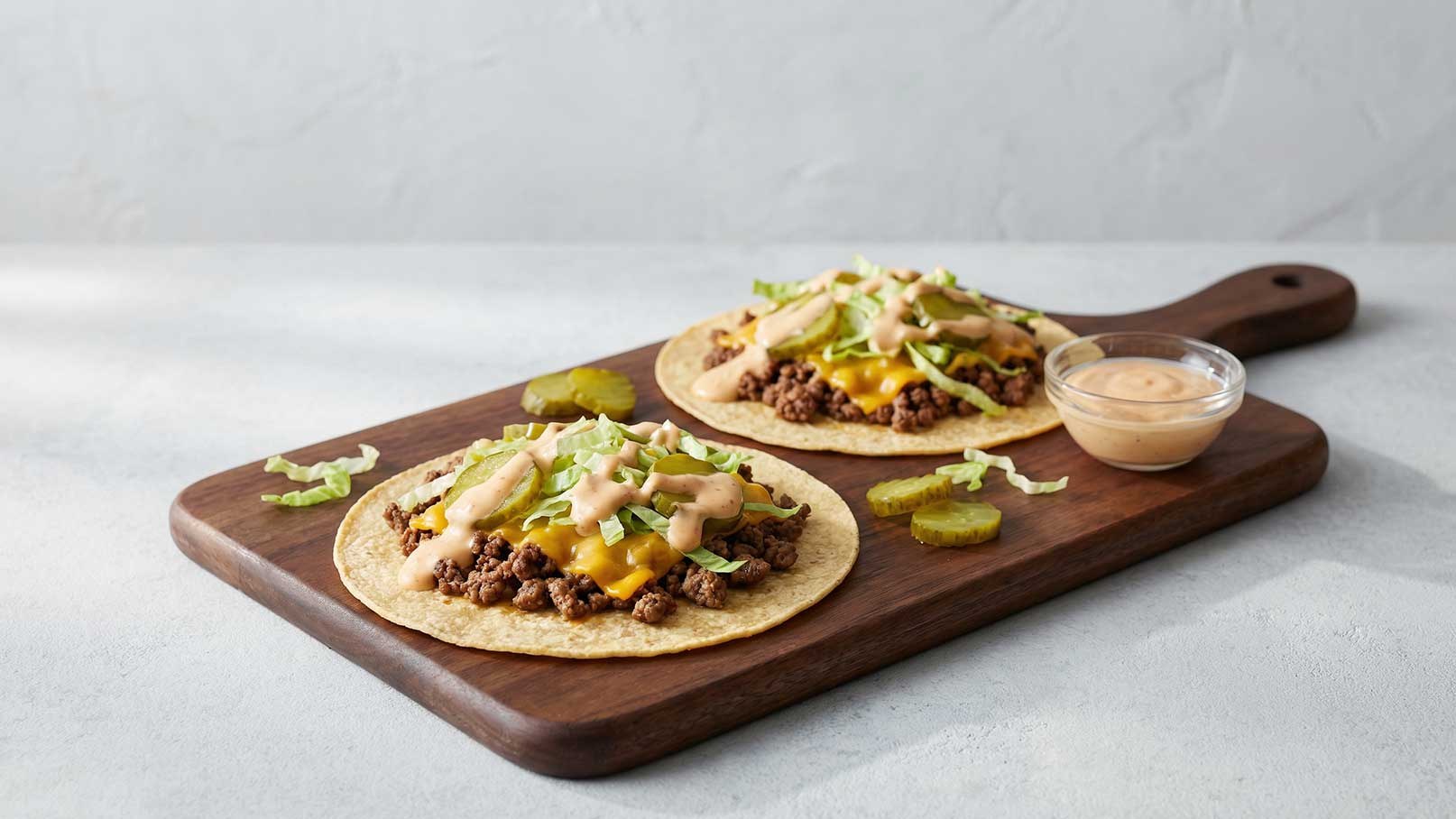 Zwei Tortillas auf einem Holzbrett, gefüllt mit gewürztem Hackfleisch, geschmolzenem Käse und Salat. Daneben befindet sich eine kleine Schüssel mit einer cremigen Sauce sowie ein paar Gurkenscheiben. Der Hintergrund ist hell und schlicht.