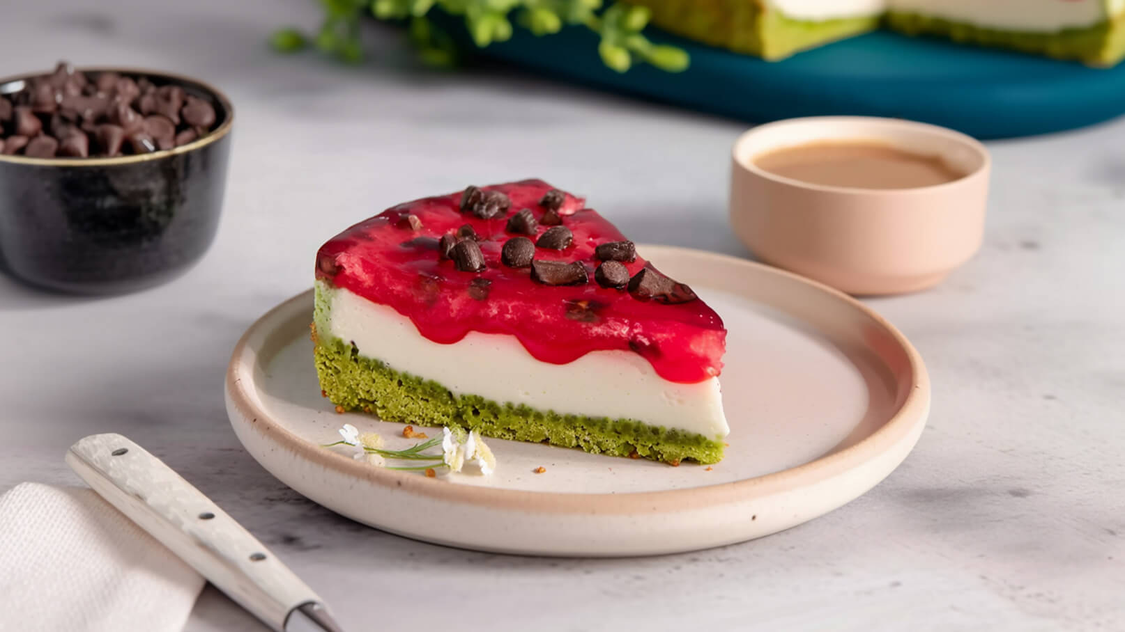 Ein Stück Käsekuchen mit grünem Boden und roter Fruchtsoße obendrauf. Die Torte ist mit Schokoladenstückchen dekoriert und auf einem weißen Teller platziert. Im Hintergrund sind eine kleine Tasse und eine Schüssel mit Schokoladenstückchen zu sehen.