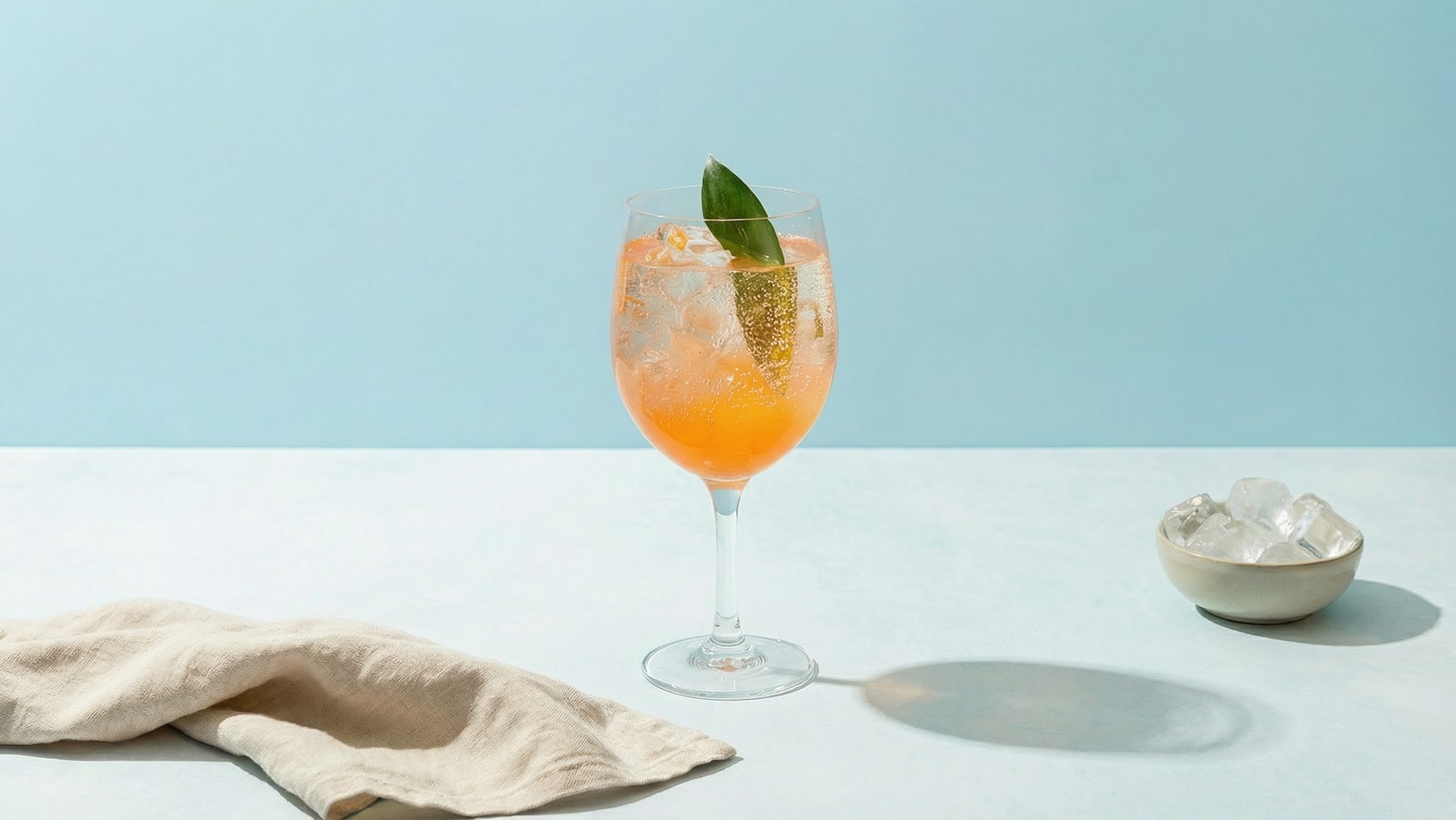 Ein frischer, orangefarbener Cocktail in einem hohen Glas, mit Eiswürfeln und einem grünen Blatt zur Dekoration. Der Hintergrund ist hellblau, während ein Becher mit weiteren Eiswürfeln daneben steht.