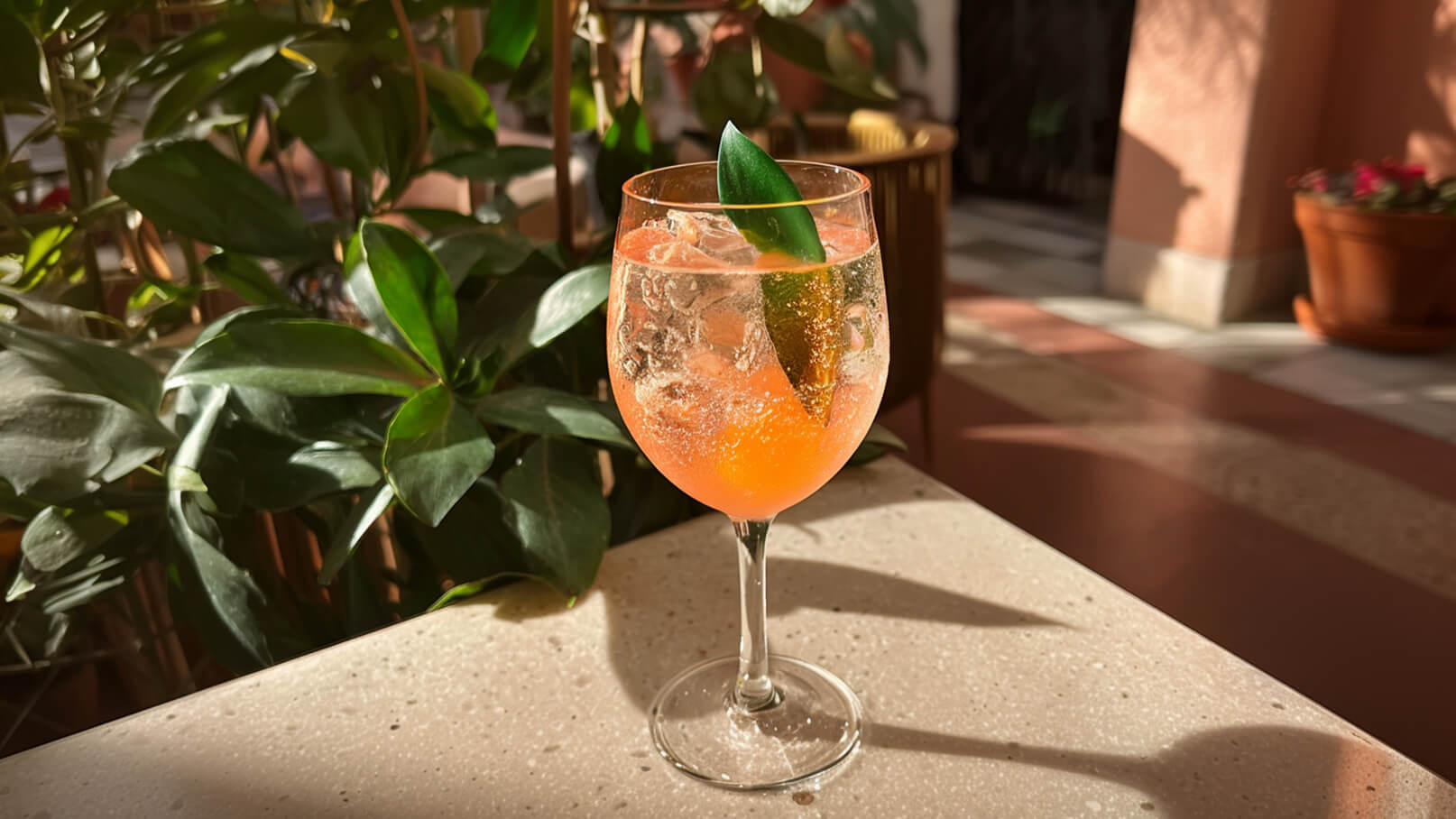 Ein Glas mit einem sprudelnden, orangefarbenen Cocktail steht auf einem Tisch. Die Oberfläche des Getränks ist mit Eiswürfeln gefüllt, und eine grüne Blattdekoration ragt heraus. Im Hintergrund sind verschwommen grüne Blätter zu sehen.