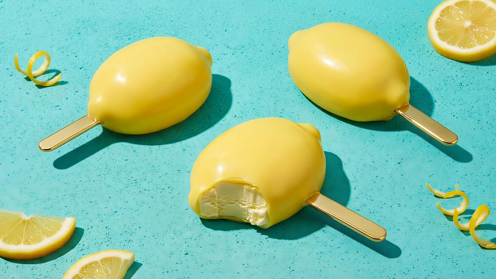 Drei gelbe Zitroneneis am Stiel liegen auf einem hellblauen Untergrund. Eines der Eis hat einen Biss, sodass die cremige Füllung sichtbar ist. Um die Eiscreme herum sind einige Zitronenscheiben und Zitronenschalen zu sehen.