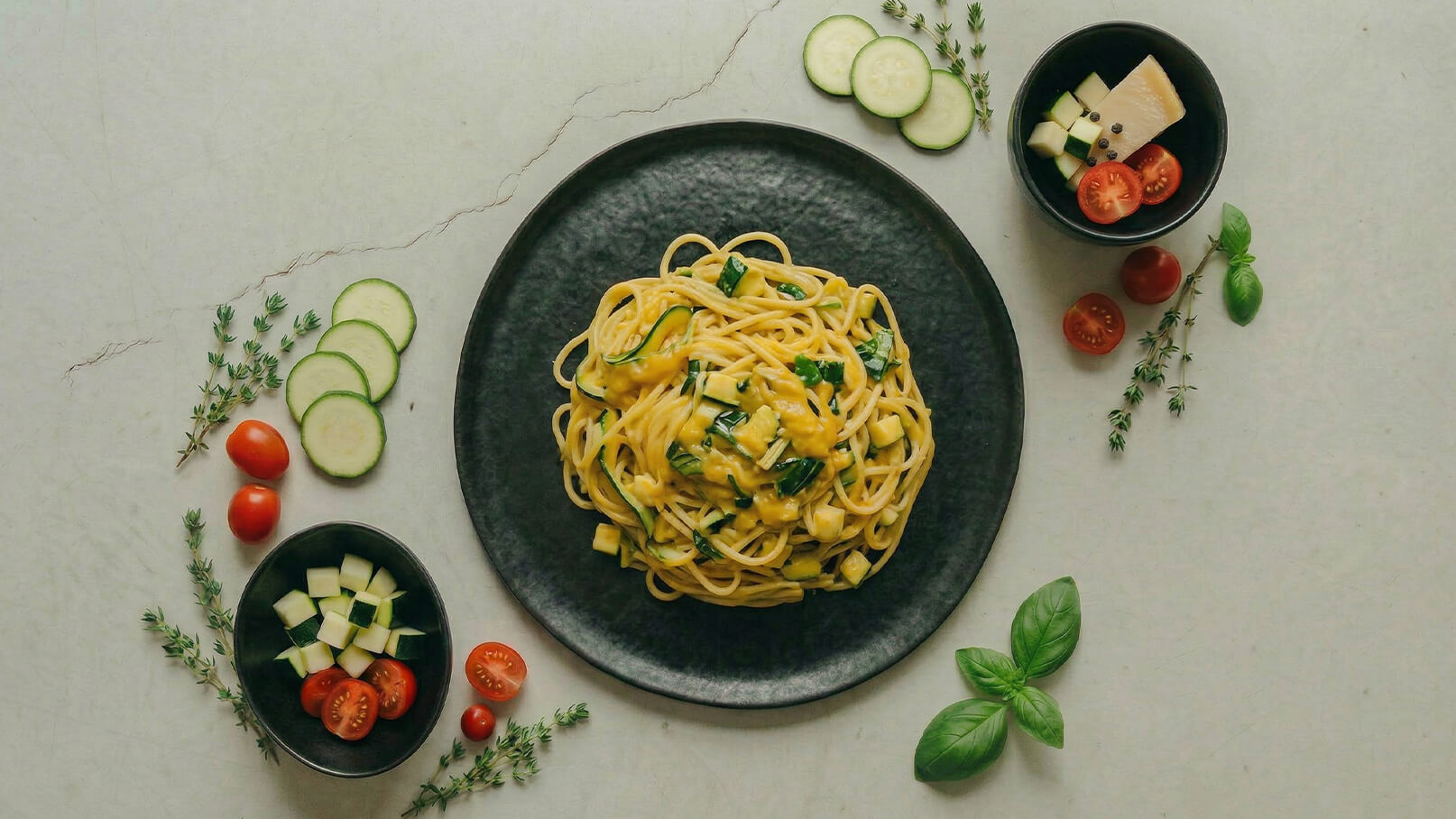 Ein schwarzer Teller mit Spaghetti, garniert mit frischen Kräutern. Daneben befinden sich kleine Schalen mit gewürfeltem Käse, Cherry-Tomaten und Gurkenscheiben, die das Gericht umrahmen. Frische Basilikumblätter und Thymianfarbe ergänzen das Bild.